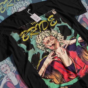 Puede incluir: Camiseta negra con un gráfico retro de una mujer con cabello rubio rizado y el texto "THE BRIDE" en amarillo. La obra de arte tiene una estética de cómic desgastada. La camiseta tiene una etiqueta que dice "LARGE".