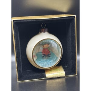Può includere: Una decorazione natalizia sferica color crema con una finestra circolare centrale che mostra una piccola figura con un cappotto rosso e un cappello verde. Il testo Merry Christmas 1978 è stampato in corsivo bianco sulla base blu interna.