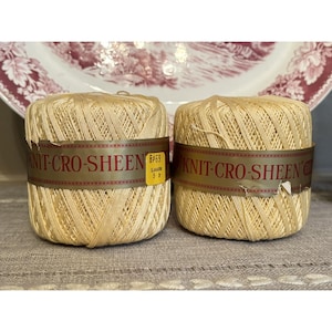 May include: Two spools of Knit-Cro-Sheen thread in a light yellow hue. Each spool is wrapped with a brown and gold label. A decorative plate with a floral design is in the background.