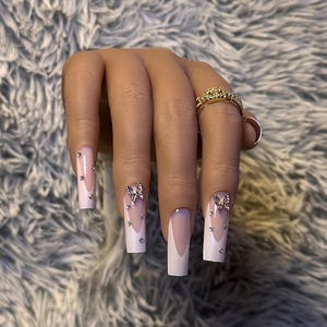 May include: Square-shaped artificial nails with a French tip design. The nails feature a light pink base with white tips, embellished with silver rhinestones and bow-shaped charms. A gold ring with a floral design is also visible.