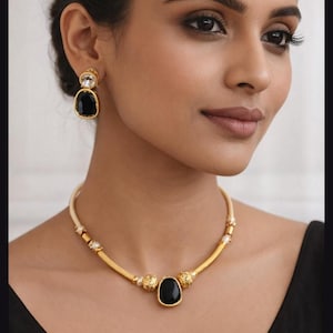 Op de afbeelding: Goudkleurige sieradenset met een ketting en oorbellen. De ketting heeft een centrale zwarte stenen hanger en een gouden ketting. De oorbellen hebben een zwarte steen en een heldere steen. De sieraden worden gedragen.