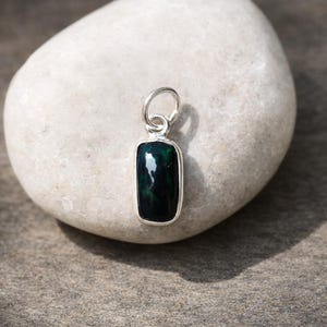 Może przedstawiać: Prostokątny wisiorek z ciemnozielonym kamieniem szlachetnym osadzonym w srebrnej ramie. Kamień szlachetny ma odblaskową powierzchnię, a wisiorek jest zawieszony na małej srebrnej pętli. Wisiorek jest prezentowany na białym kamieniu.
