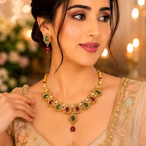 May include: A gold-toned necklace and matching stud earrings set featuring alternating teardrop-shaped red and green stones. The necklace includes small white pearls between the stones, with a central teardrop pendant and a dangling red bead.