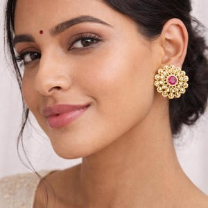 Könnte beinhalten: Eine Frau trägt goldfarbene, blütenförmige Ohrringe mit einem roten Edelstein in der Mitte. Die Ohrringe sind mit kleinen, umlaufenden Verzierungen versehen. Auf der Stirn der Frau ist ein kleiner roter Punkt zu sehen.