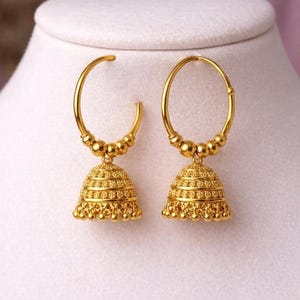 May include: A pair of gold-colored hoop earrings with a bell-shaped charm. The earrings have a detailed, textured design and are displayed on a white surface against a soft pink background. The earrings are a classic jewelry item.