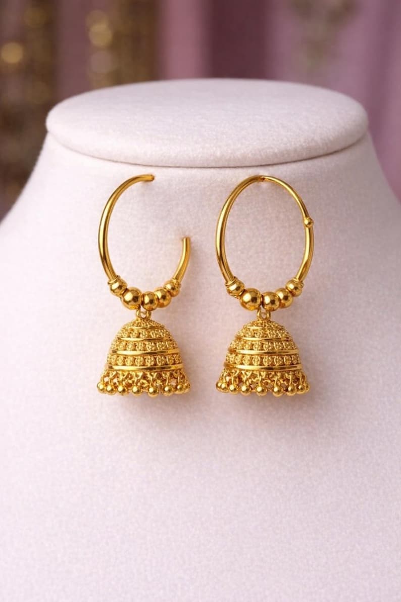 May include: A pair of gold-colored hoop earrings with a bell-shaped charm. The earrings have a detailed, textured design and are displayed on a white surface against a soft pink background. The earrings are a classic jewelry item.