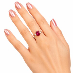 May include: A gold ring featuring a central emerald-cut red gemstone flanked by two smaller, square-cut clear stones. The band is thin and polished, showcasing a classic three-stone design that sits prominently on the finger with a vibrant red hue.