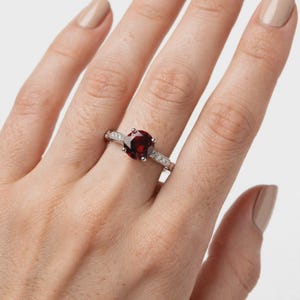 Peut inclure: Une bague en argent ornée d'une grande pierre précieuse grenat ronde d'un rouge profond au centre. L'anneau est fabriqué en argent poli et est agrémenté de petites pierres claires et scintillantes incrustées le long de la tige.