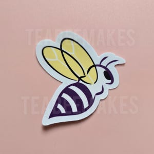 May include: A sticker featuring a stylized bee design. The bee has a white body with purple stripes, yellow wings, and black outlines. The sticker is on a pink background.
