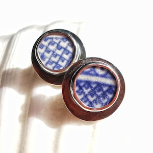 Könnte beinhalten: Ein Paar runde Ohrstecker in Silberoptik. Jeder Ohrring hat einen Einsatz aus Keramik mit blau-weißem Muster. Die Ohrringe sind vor einem weißen, strukturierten Hintergrund platziert, wobei sich das Licht im Metall spiegelt.