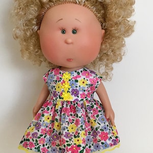 May include: A doll with a round face, green eyes, and blonde curly hair. The doll wears a floral print dress with pink, yellow, and purple flowers. The dress has a yellow trim and a yellow lace detail.
