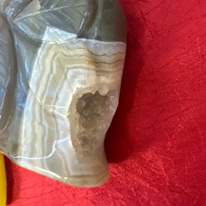 Pode incluir: Uma escultura em pedra esculpida de uma folha com um interior semelhante a uma geodo. A escultura é principalmente cinza com faixas brancas e bege, e uma seção escavada revela uma cavidade cheia de cristais. O fundo é vermelho.