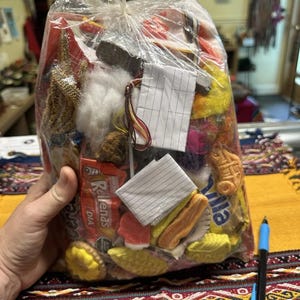 Puede incluir: Una bolsa de plástico transparente llena de diversos materiales de artesanía coloridos, incluyendo retazos de tela, caramelos y pequeños objetos decorativos. Una mano sostiene la bolsa, y un bolígrafo descansa sobre un textil estampado.