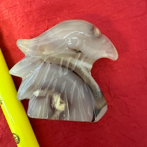 Pode incluir: Uma escultura em pedra esculpida da cabeça de uma águia, com tons de cinza, branco e castanho. A escultura está sobre um fundo vermelho. A cabeça da águia é detalhada com penas esculpidas e um bico afiado.