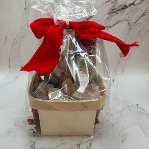 Peut inclure: Un panier cadeau enveloppé dans du cellophane transparent et noué avec un grand nœud en velours rouge. Le panier est en bois clair et contient divers articles, y compris des éléments décoratifs.
