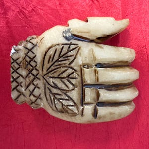 Puede incluir: Un objeto tallado en forma de mano, de color blanquecino, con intrincados diseños de hojas y sombreado cruzado. Los dedos están claramente definidos y el objeto tiene un aspecto ligeramente envejecido. El fondo es rojo liso.