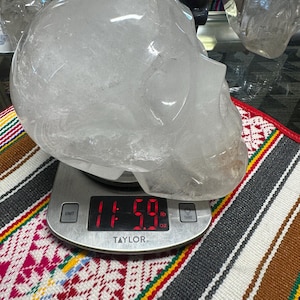 Op de afbeelding: Een heldere kwartskristallen schedel wordt gewogen op een digitale Taylor-weegschaal, met een gewicht van 5,26 kg. De schedel is geplaatst op een kleurrijk patroontextiel. Andere kristallen schedels zijn zichtbaar op de achtergrond.