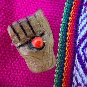 Pode incluir: Um broche de madeira esculpida em forma de punho com um olho de contas vermelho. O broche é colocado sobre um tecido rosa vibrante, com uma borda listrada colorida em verde, vermelho, laranja e roxo.