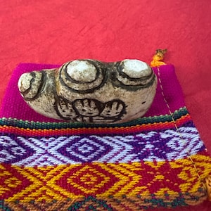 Pode incluir: Um artefato de pedra esculpida com desenhos circulares repousa sobre um tecido vibrante e tecido. O tecido apresenta um padrão geométrico colorido em roxo, amarelo, vermelho e verde. O fundo é vermelho liso.