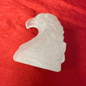 Puede incluir: Una escultura de cabeza de águila de cristal tallado transparente. El águila está representada de perfil, con plumas detalladas y un pico afilado. La escultura está sobre un fondo rojo.