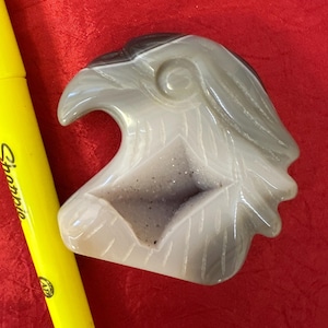 Peut inclure: Une sculpture en pierre taillée représentant une tête d'aigle dans des tons de gris et de blanc, avec un intérieur sombre. La sculpture est posée sur un fond rouge. Un marqueur Sharpie jaune est dans l'image.