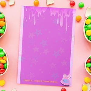 Puede incluir: Un fondo rosa con varios recipientes en forma de huevo llenos de caramelos de colores. También hay un cuaderno morado con un diseño de goteo y texto. Los caramelos y trozos de chocolate dispersos completan la escena.