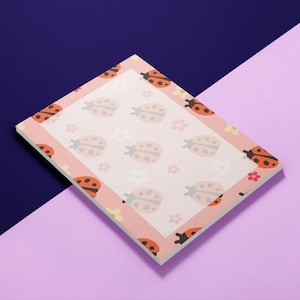 May include: A rectangular notepad with a pink border and a white interior. The border is decorated with orange ladybugs and small flowers. The interior features a repeating pattern of ladybugs and flowers. The notepad is angled on a purple and blue background.