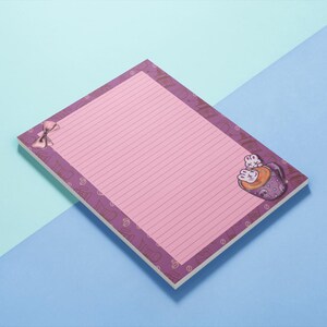 May include: A lined notepad with a pink interior and a purple border. The border features a repeating pattern of hearts and a bow. A cartoon illustration of two rabbits in a cup is in the corner. The notepad is on a blue and teal background.