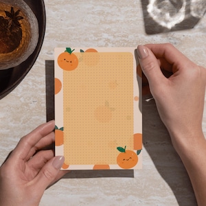 May include: A notepad with an orange theme, held by a person. The notepad has a light orange background with small dots and orange illustrations of smiling oranges with green leaves. The notepad's border is decorated with orange circles.