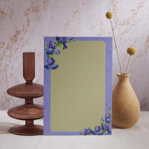 May include: A rectangular notepad with a light green interior and a lavender border adorned with watercolor irises. A brown glass candle holder and a small vase with dried flowers are on either side of the notepad.