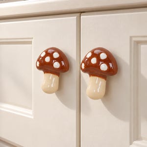 Puede incluir: Dos pomos de armario de cerámica con forma de seta, con sombreros marrones brillantes con puntos blancos y tallos lisos de color crema. Estos herrajes decorativos están montados en la parte frontal de dos puertas de armario color crema.