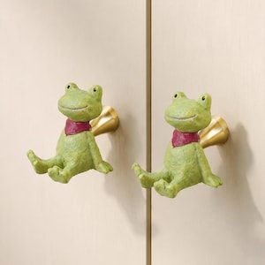Op de afbeelding: Twee kastknoppen in de vorm van zittende groene kikkers met een textuurhuid en kleine rode halsdoekjes. Elke kikkerfiguur is bevestigd aan een conische, goudkleurige metalen voet, ontworpen om op een lichtgekleurde houten kastdeur te worden gemonteerd.