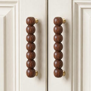 May include: Two vertical cabinet pulls featuring a series of seven dark brown wooden spheres stacked in a column. Each handle is secured to the off-white cabinet doors with small, circular brass-toned metal hardware at the top and bottom ends.