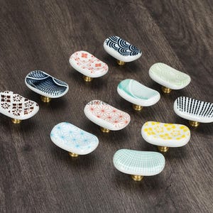 May include: A selection of ceramic cabinet knobs with diverse patterns and colours. The knobs have a white base with designs in blue, red, yellow, and green. Each knob has a brass-coloured base and is set against a dark wood surface.