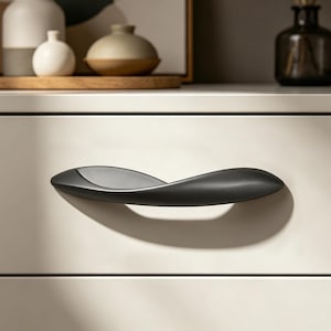 May include: A close-up of a black drawer pull with a unique, curved design. The handle is mounted on a white drawer, with a shadow cast below. The pull has a smooth, matte finish, and is a modern accent for furniture.