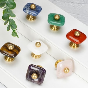 May include: A collection of seven square cabinet knobs featuring polished natural stone tops in blue, green, red, brown, white, purple, and light pink, each accented with a central brass-toned metal stud and a matching cylindrical brass base.