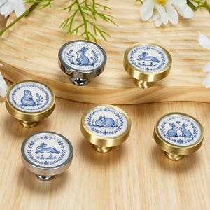 May include: A set of six round cabinet knobs featuring blue and white ceramic inserts with various rabbit illustrations. The knobs have metallic bases in gold and silver tones, each decorated with a blue floral wreath border around the rabbit.