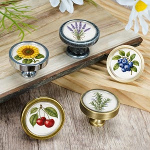 May include: Five round cabinet knobs featuring botanical illustrations on white circular inserts. The designs include a yellow sunflower, purple lavender, blue blueberries, red cherries, and green rosemary, set in metallic silver, gold, and dark gray frames.