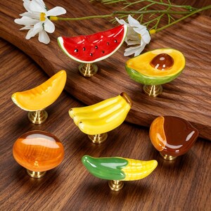 May include: A collection of seven ceramic cabinet knobs shaped like various fruits and vegetables, including a watermelon slice, avocado half, banana bunch, corn cob, and other rounded fruit forms, each mounted on a small brass-colored metal base.