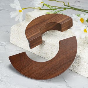 May include: Two dark brown wooden crescent-shaped pieces with a smooth, polished finish and visible wood grain. The curved segments are arranged to form a circular shape, showcasing the natural texture and deep tones of the wood material.