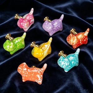 Peut inclure: Un ensemble de sept boutons de placard en forme de chat avec une finition en résine marbrée rose, violette, rouge, verte, jaune, turquoise et orange. Chaque bouton possède une base en métal doré et une petite queue dressée.