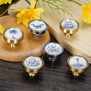 May include: A set of six round cabinet knobs with a metallic gold finish, each featuring a unique blue and white nautical illustration including a palm tree, a whale, a sailing ship, an octopus, seashells, and a lighthouse on the circular face.