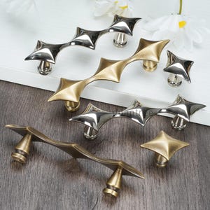 以下が含まれることがあります： さまざまな金属仕上げの装飾的なキャビネット金具の品揃え。コレクションには、幾何学的なデザインの星型のノブとハンドルが含まれています。金具は木製の表面に表示されています。