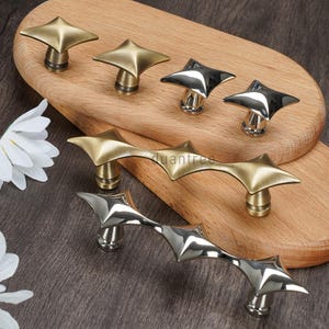 May include: A collection of star-shaped cabinet knobs and pulls in brass and silver finishes. The knobs have a four-point star design, while the pulls feature a series of connected stars. The hardware is displayed on a wooden tray.