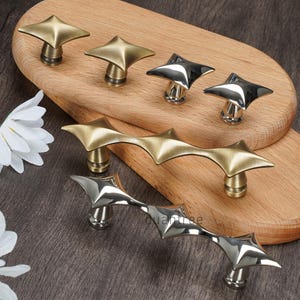 May include: A collection of star-shaped cabinet hardware in brass and silver finishes. The hardware includes individual knobs and longer pulls, all with a geometric, starburst design. The hardware is displayed on a wooden surface.