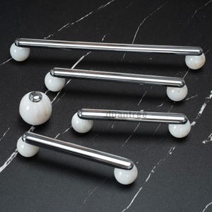 May include: A collection of cabinet pulls with a polished chrome finish and white marble sphere accents. The pulls are rectangular with rounded ends and are arranged on a black and white marble surface. The text "duantree" is visible on one of the pulls.