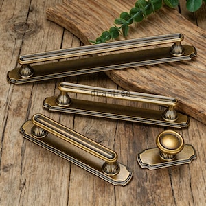 May include: An assortment of antique brass cabinet hardware, including long pull handles and a round knob with a rectangular backplate. The hardware features a detailed, ornate design and a warm, aged finish. The pieces are arranged on a rustic wooden surface.
