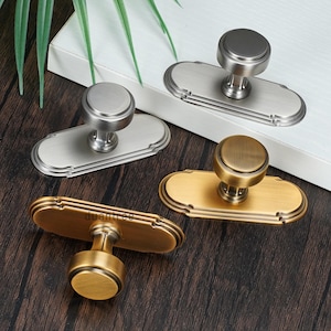 May include: Four cabinet pulls in brushed nickel and antique brass finishes. Each pull has a round knob on an oval backplate with decorative edging. They are arranged on a dark wood surface.