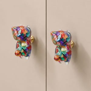 May include: Two cat-shaped cabinet knobs made of clear resin filled with a variety of colourful, polished stone and shell fragments. Each knob features a gold-toned metal base that attaches to the surface, showcasing a vibrant, multi-coloured mosaic.