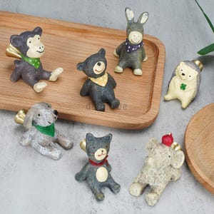 May include: A collection of small, animal-shaped figurines, including a monkey, bear, donkey, hedgehog, dog, cat, and elephant. Each figurine is painted with details like bandanas and hats. The figurines are arranged on a wooden tray.